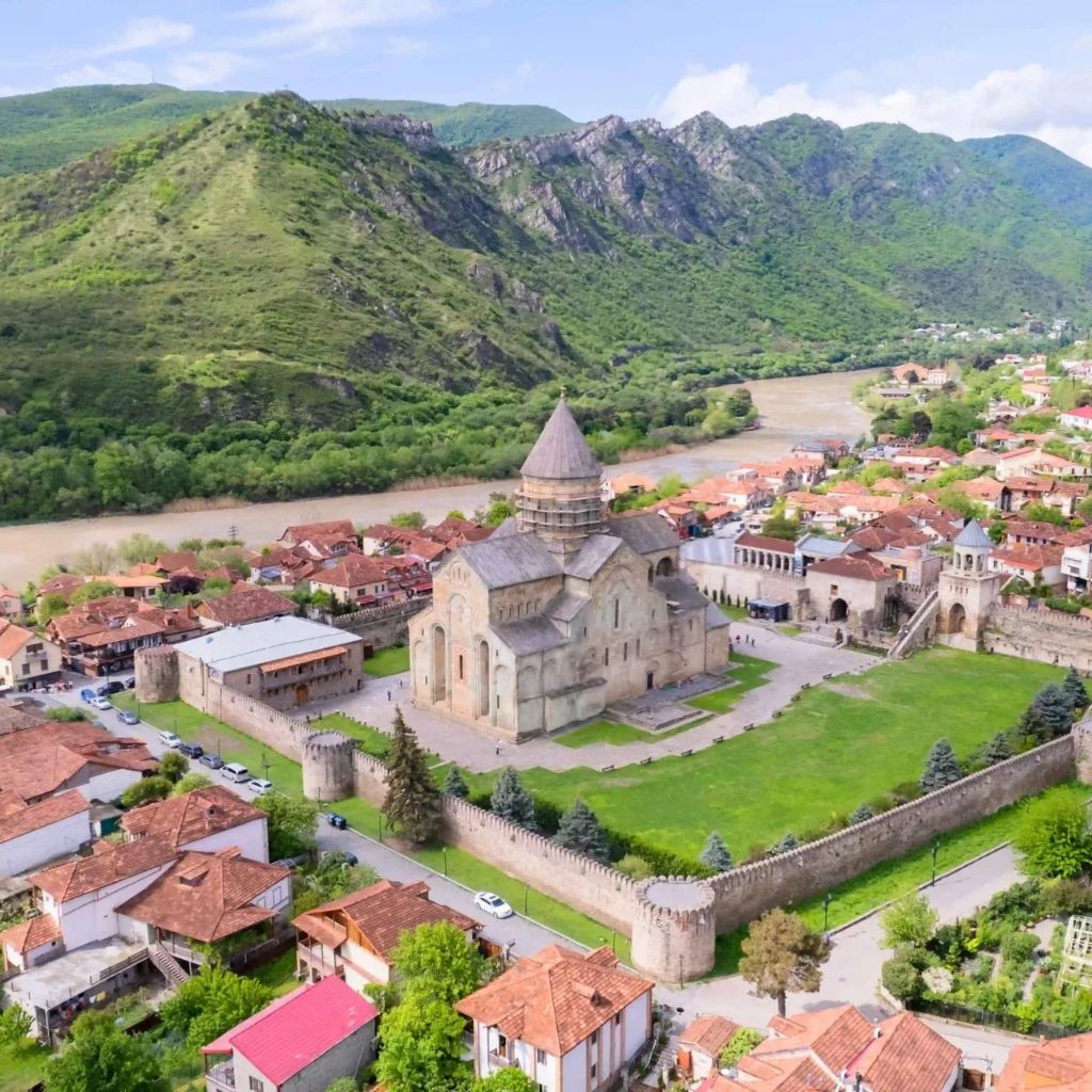 Aerial view of Mtskheta Georgia historic stone church with a conical roof, surrounded by fortified walls. Nearby red-roofed houses and a river, set against lush green hills.