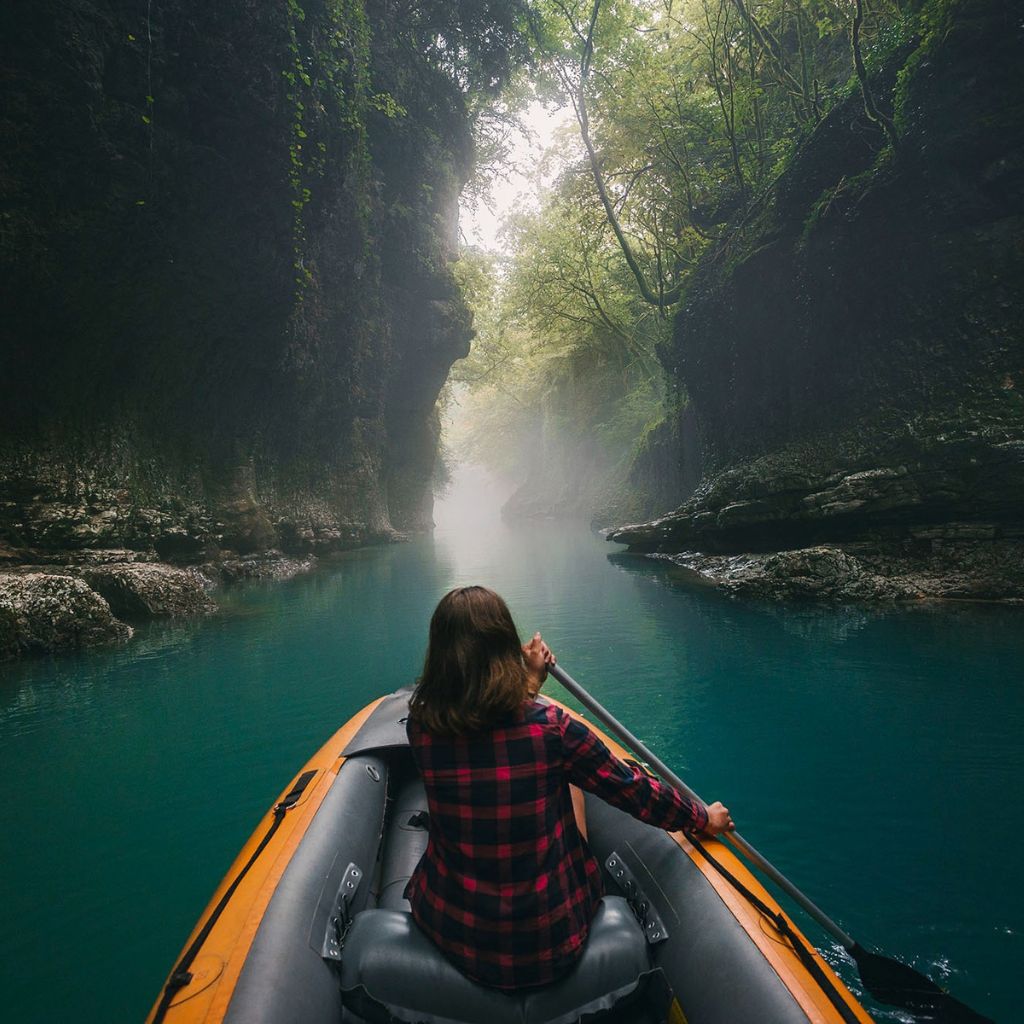 A person in a plaid shirt paddles an inflatable boat through a narrow, misty canyon. The water is calm and turquoise, lined with lush green trees and rocky cliffs.