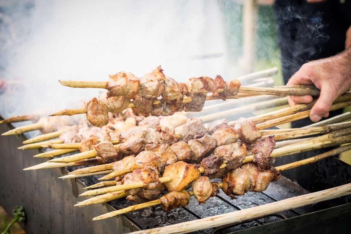 Mtswadi Georgian Food Grilled meat skewers sizzling over an outdoor barbecue, with smoke rising. A hand adjusts the sticks, evoking a sense of summer and savory aroma.