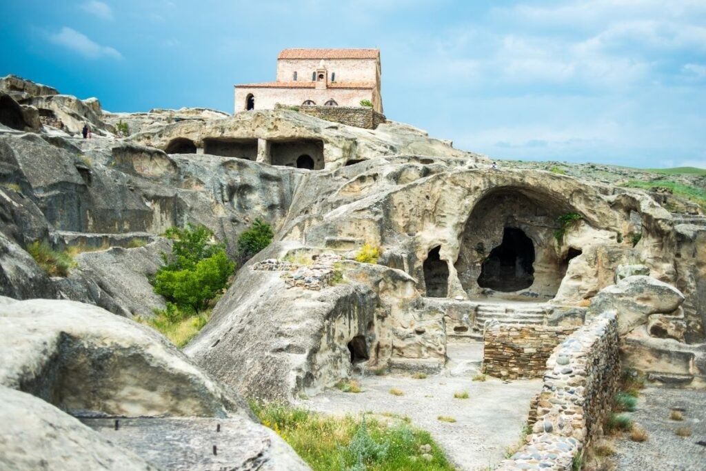 Ancient stone Uplistsikhe cityscape featuring a cave dwelling below a rustic church on a hill. The sky is cloudy, evoking a sense of history and mystery.