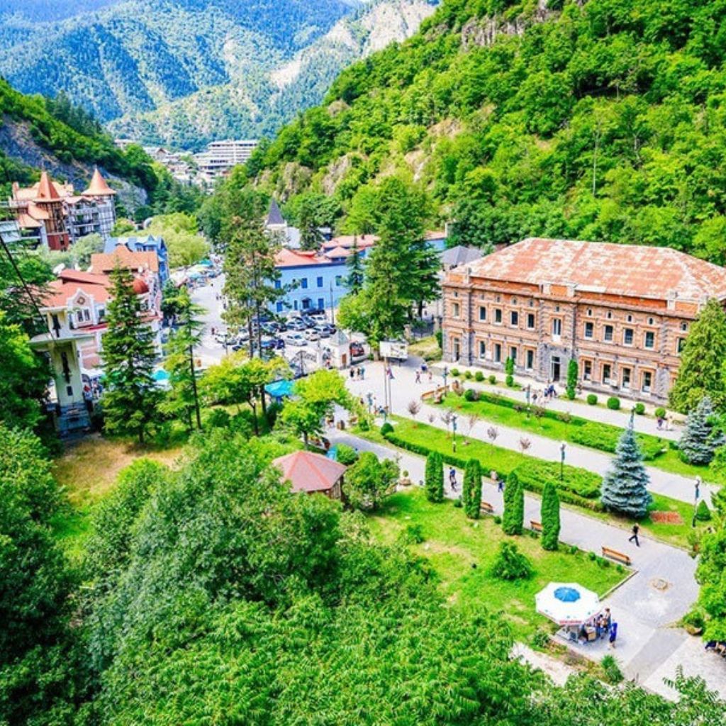 A vibrant aerial view of a scenic town Borjomi Georgi nestled in lush green mountains. Historic buildings, people strolling, and colorful umbrellas evoke a peaceful, lively atmosphere.