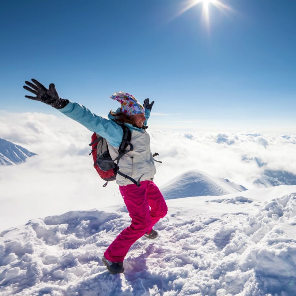 A person joyfully leaps in the Gadurui Georgia snowy mountains under a bright sun. They wear colorful winter gear, conveying excitement and freedom, with clouds below.