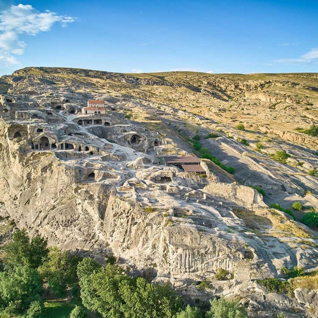 Aerial view of ancient rock-cut dwellings on a rugged cliff, under a clear blue sky. Trees line the base, adding contrast to the arid landscape.