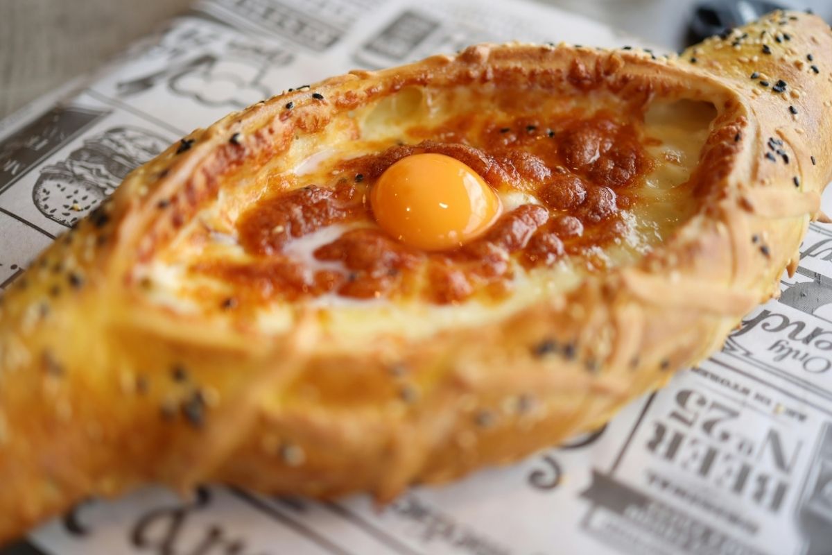 Kachapuri Food : Oval-shaped bread with a golden crust, topped with melted cheese, and a soft yolk egg in the center. Placed on newspaper, conveying warmth.