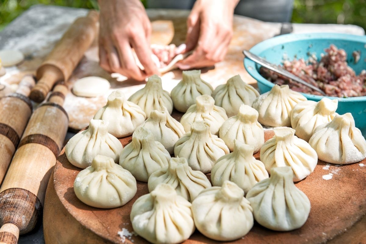 Person shaping Georgian food khinkali dumplings by hand on a wooden board, with uncooked dumplings, rolling pins, and a bowl of filling visible.